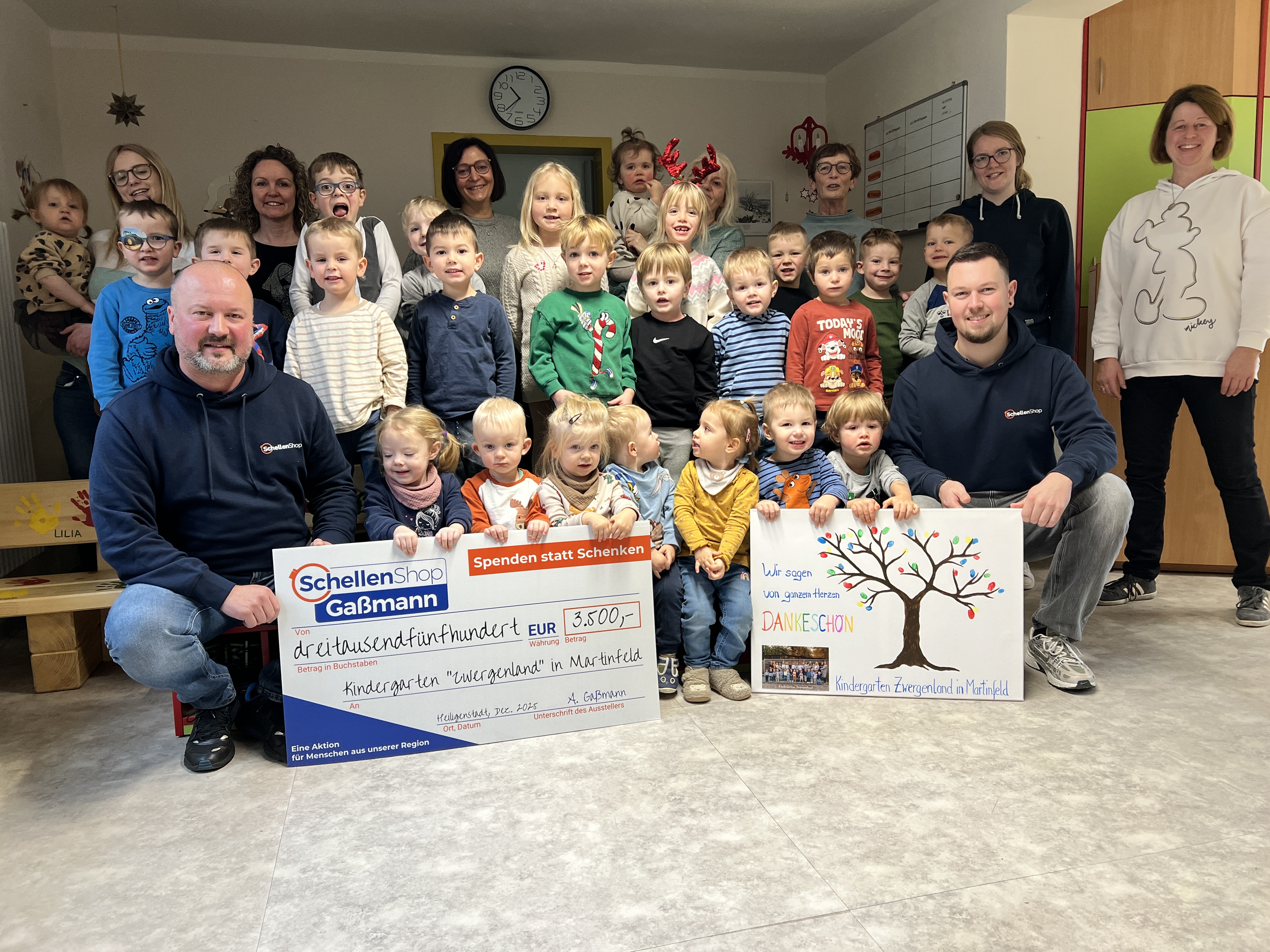 Eine große Gruppe von lachenden Kindergartenkindern und Erwachsenen posiert stolz im Kindergarten Zwergenland in Martinfeld. Zwei Erwachsene halten einen großen Scheck über 3.500 Euro von SchellenShop Gaßmann und ein Dankeschön-Plakat mit bunten Handabdrü
