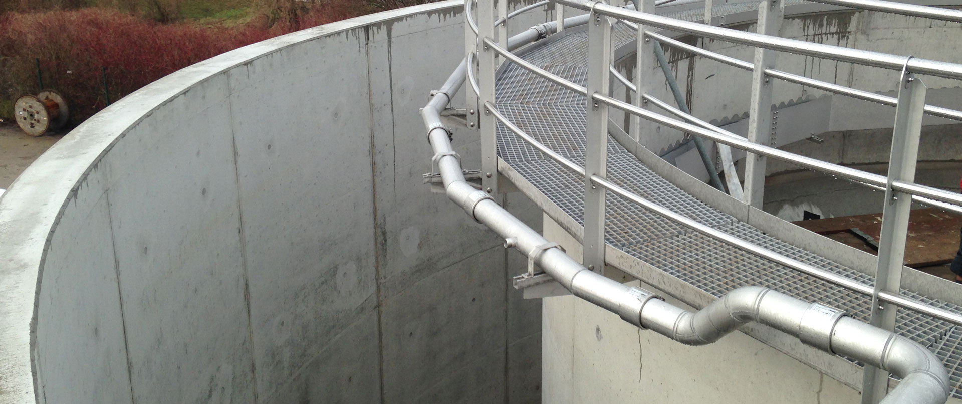 Ein Rohrleitungssystem mit Rohrkupplungen verläuft entlang einer runden Betonwand unterhalb eines Metallstegs in einer Industrieanlage.