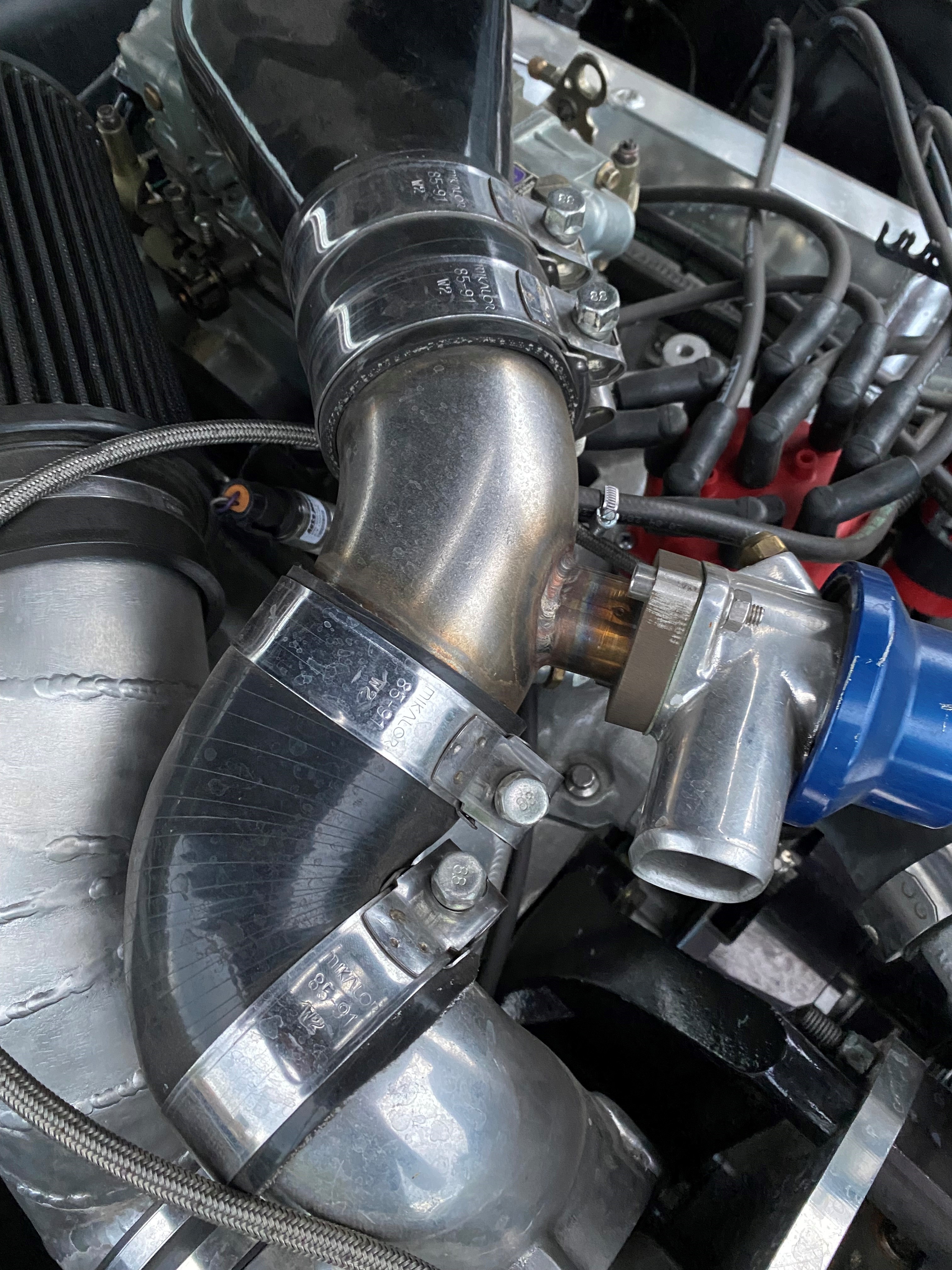 Vier Gelenkbolzenschellen befestigen schwarze Gummischläuche an silbernen Metallrohren im Motorraum eines Fahrzeugs. Die glänzenden Schellen sorgen für eine sichere Verbindung der Komponenten.