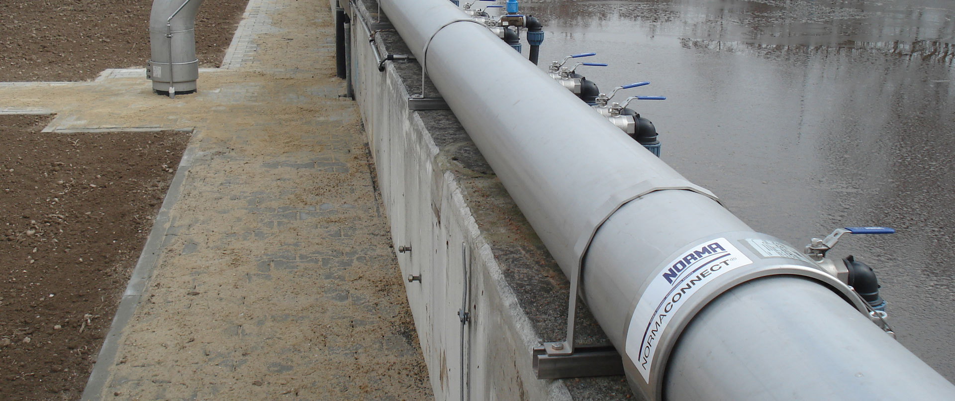 Graues Industrierohr mit mehreren blauen Ventilen und 'NORMA CONNECT' Rohrkupplungen, montiert auf einer Betonmauer, mit feuchtem Boden und reflektierendem Wasser im Hintergrund.