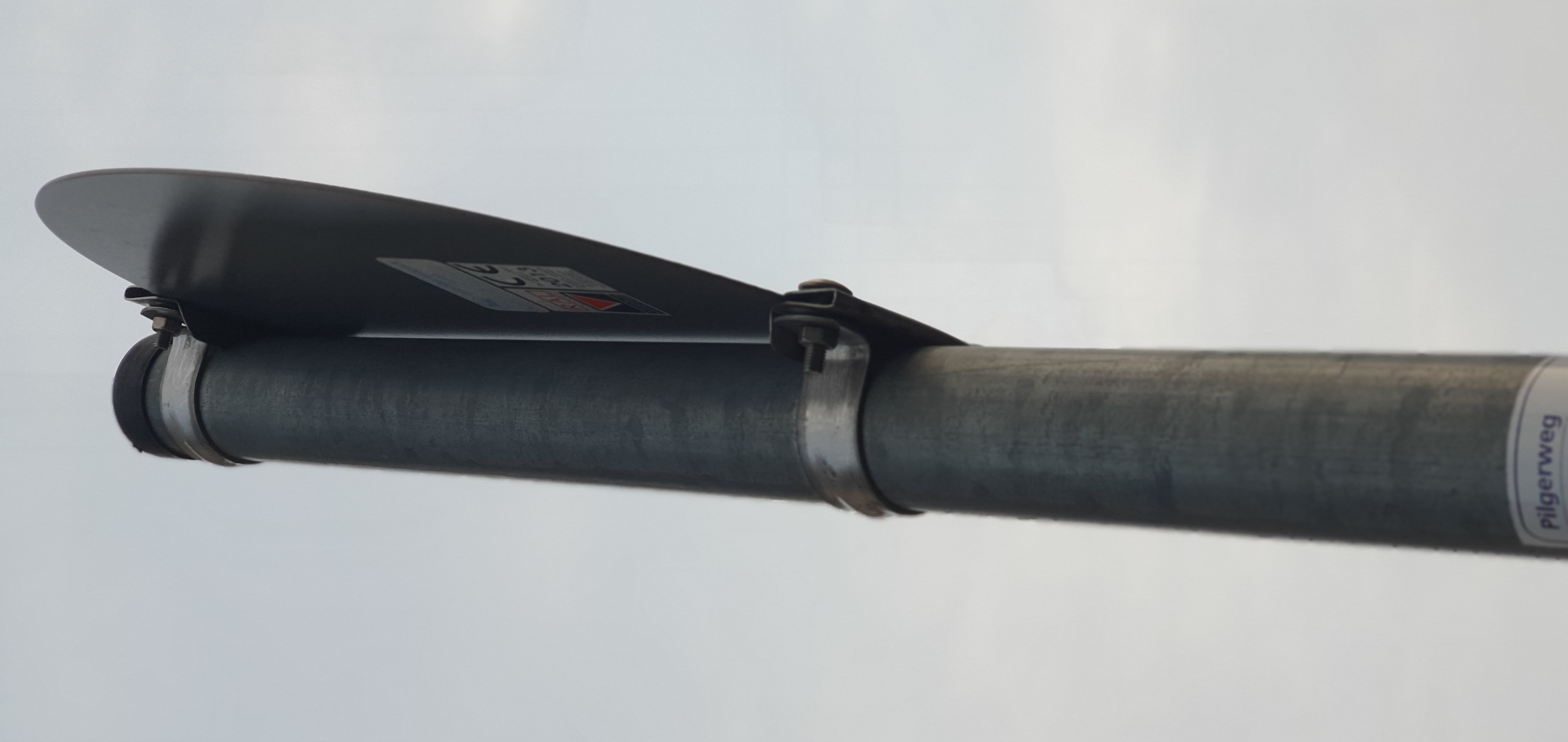 Ein grauer Metallpfosten mit einem dunklen, gekrümmten Schild, das mit zwei Metallbändern befestigt ist. Auf dem Schild ist ein Aufkleber mit der Aufschrift 'CE 2013' zu erkennen. Am unteren Ende des Pfostens ist ein weißes Etikett mit dem Schriftzug 'Pil