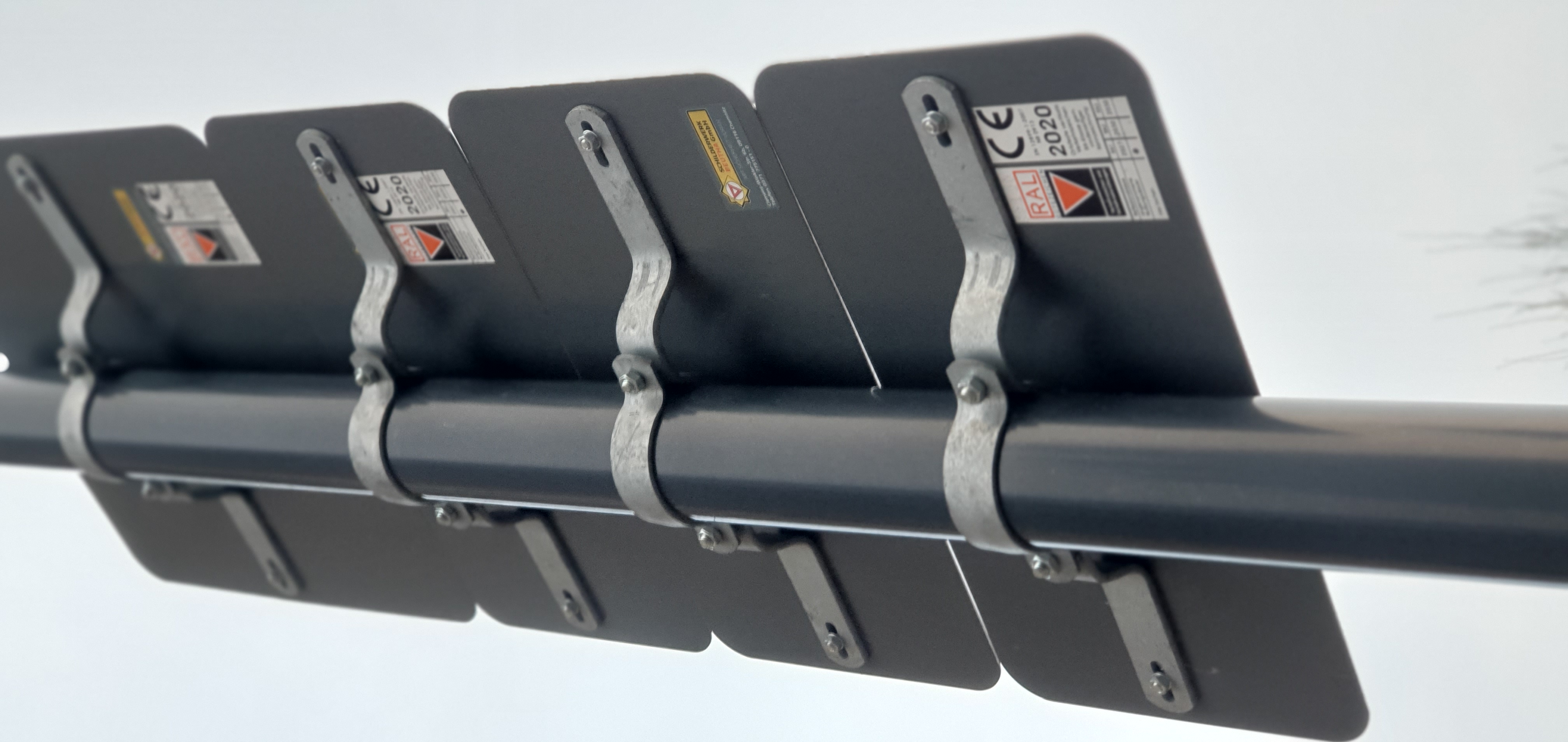 Rückansicht von mehreren vertikal gestapelten, dunklen Verkehrszeichen, die mit silbernen Halterungen an einem grauen Mast befestigt sind. Auf einigen Schildern sind Produktetiketten wie RAL und CE 2020 sichtbar.