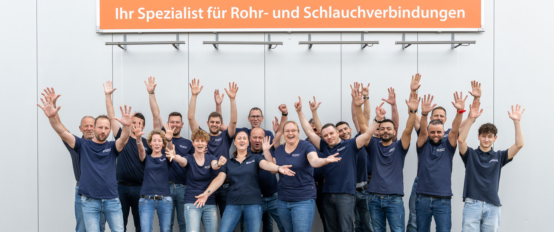 Unser engagiertes Team – Ihr Spezialist für Rohr- und Schlauchverbindungen. Lachende Mitarbeiter in blauen Firmen-T-Shirts winken vor einem Gebäude mit dem orangefarbenen Schild 'Ihr Spezialist für Rohr- und Schlauchverbindungen'.