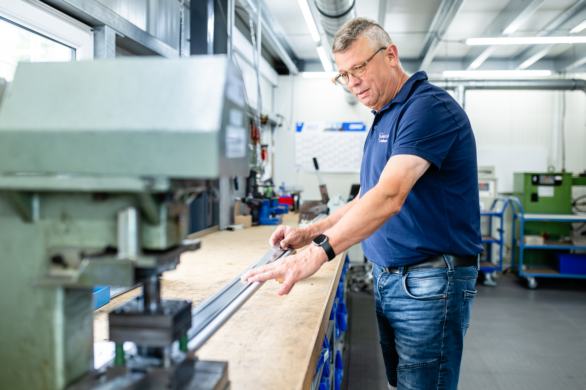 Mitarbeiter bei der Qualitätskontrolle und Bearbeitung eines Metallprofils in der Werkstatt. Ein Mann in blauem Polohemd mit Brille misst ein langes Metallprofil präzise an einer Werkbank in einer modernen Fertigungshalle.