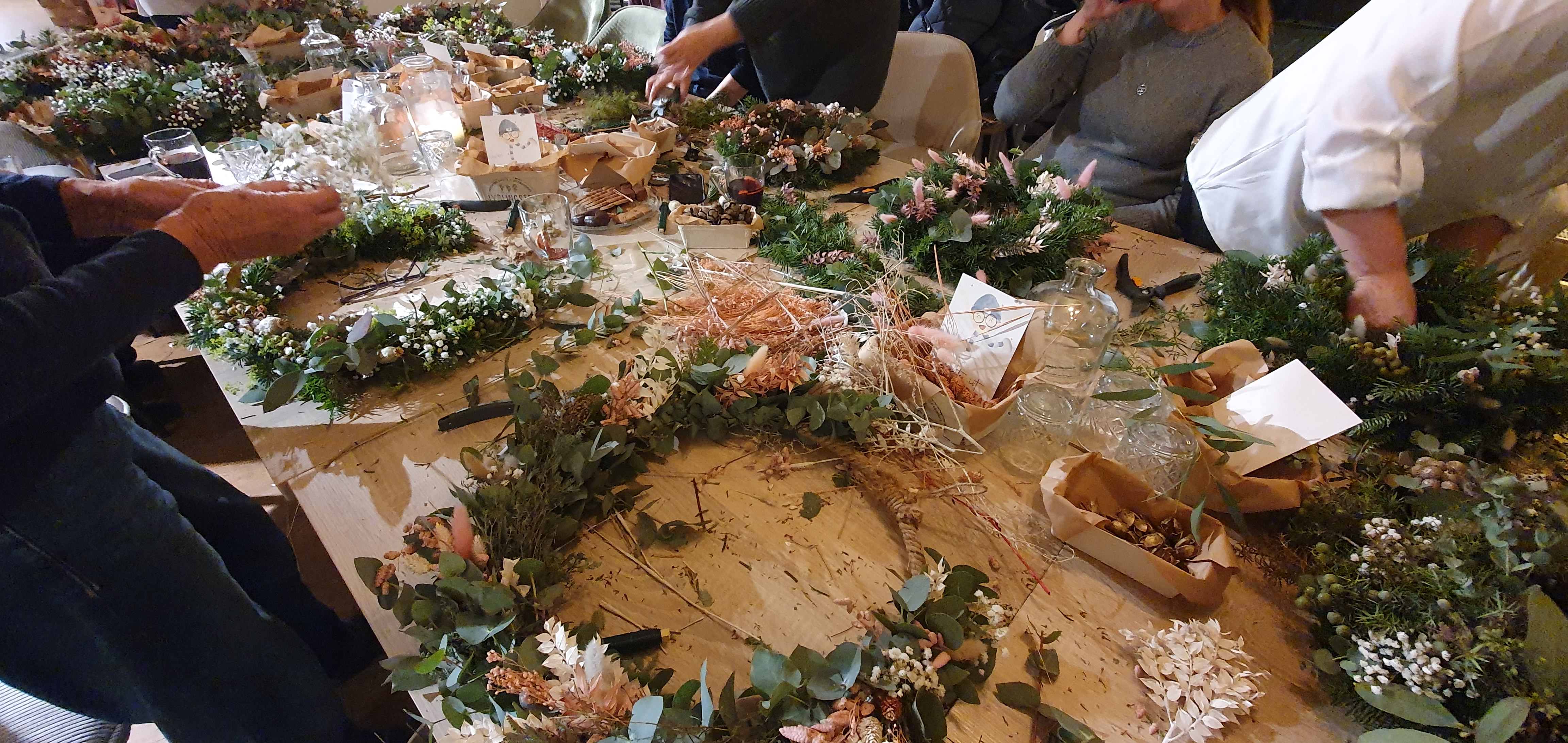 Mehrere Personen basteln gemeinsam festliche Weihnachtskränze aus Tannengrün, Eukalyptus und Trockenblumen an einem Holztisch. Dies ist das kreative Teambuilding-Event 'Zauberhafte Vorweihnachtszeit: Unser kreatives Teambuilding bei Ly’s'.